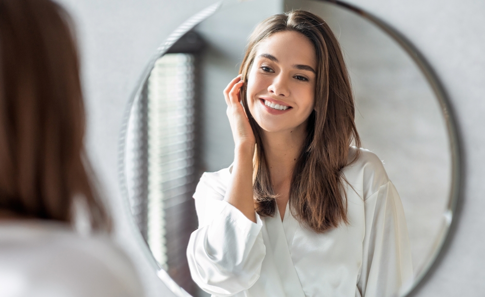 Woman looking in the mirror smiling after laser hair removal for PCOS in NYC